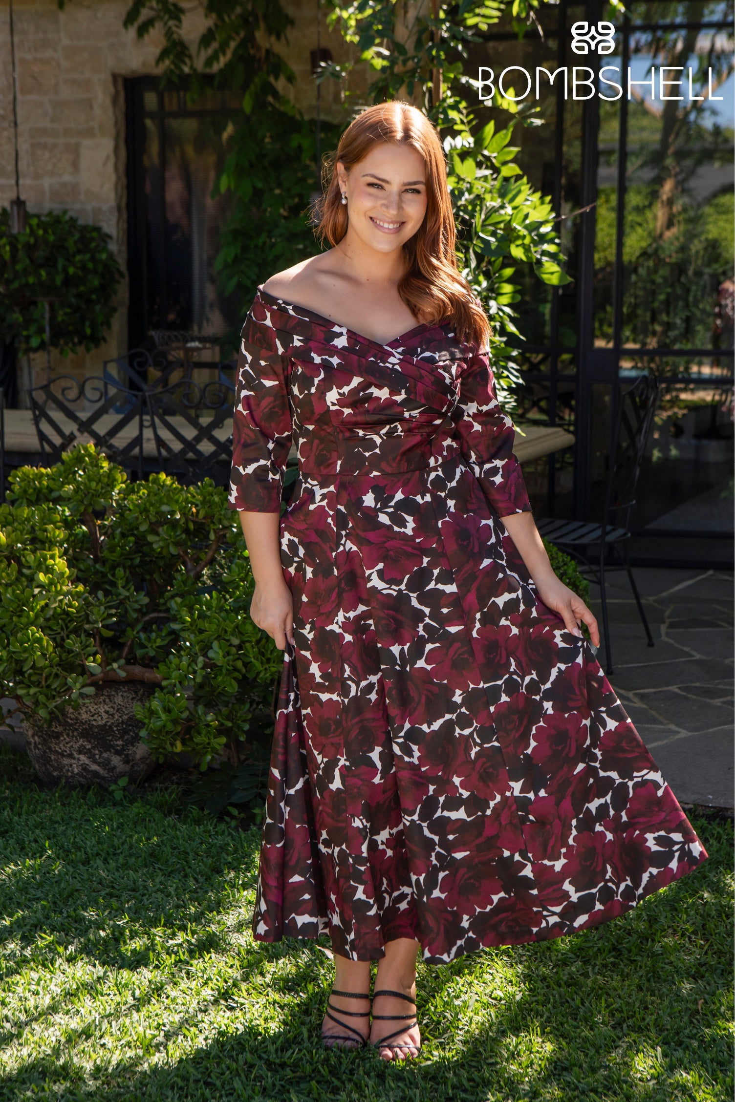 Woman in a floral dress standing outdoors with greenery and a building in the background.
