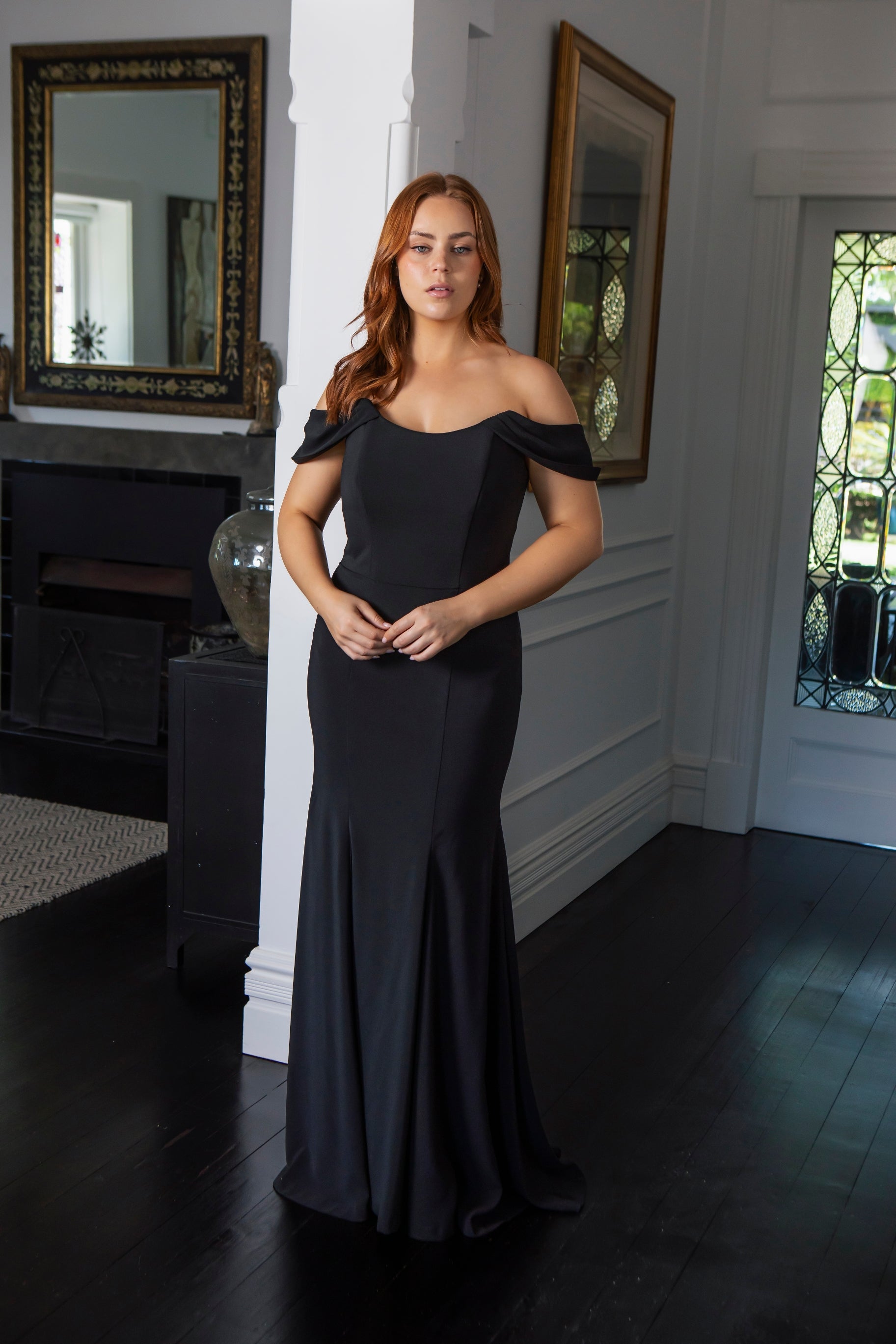 Woman in a black off-shoulder gown standing in a room with mirrors and a fireplace.