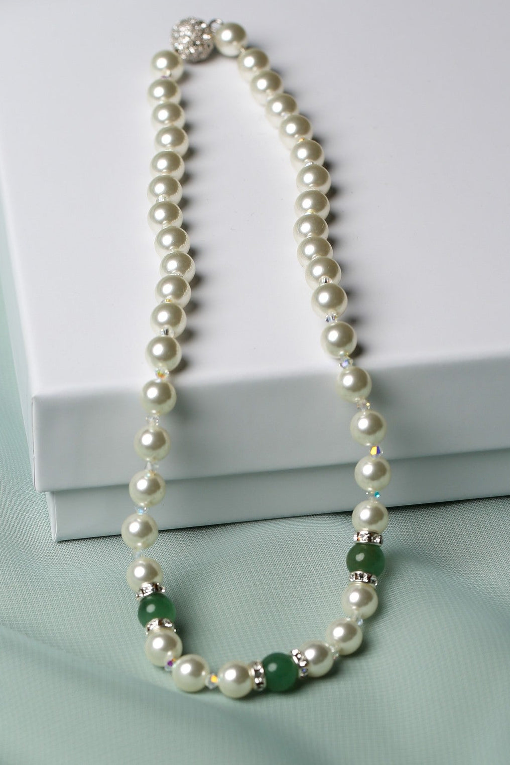 A pearl necklace with small green and silver accent beads, draped elegantly over a white gift box on a soft mint green fabric background.