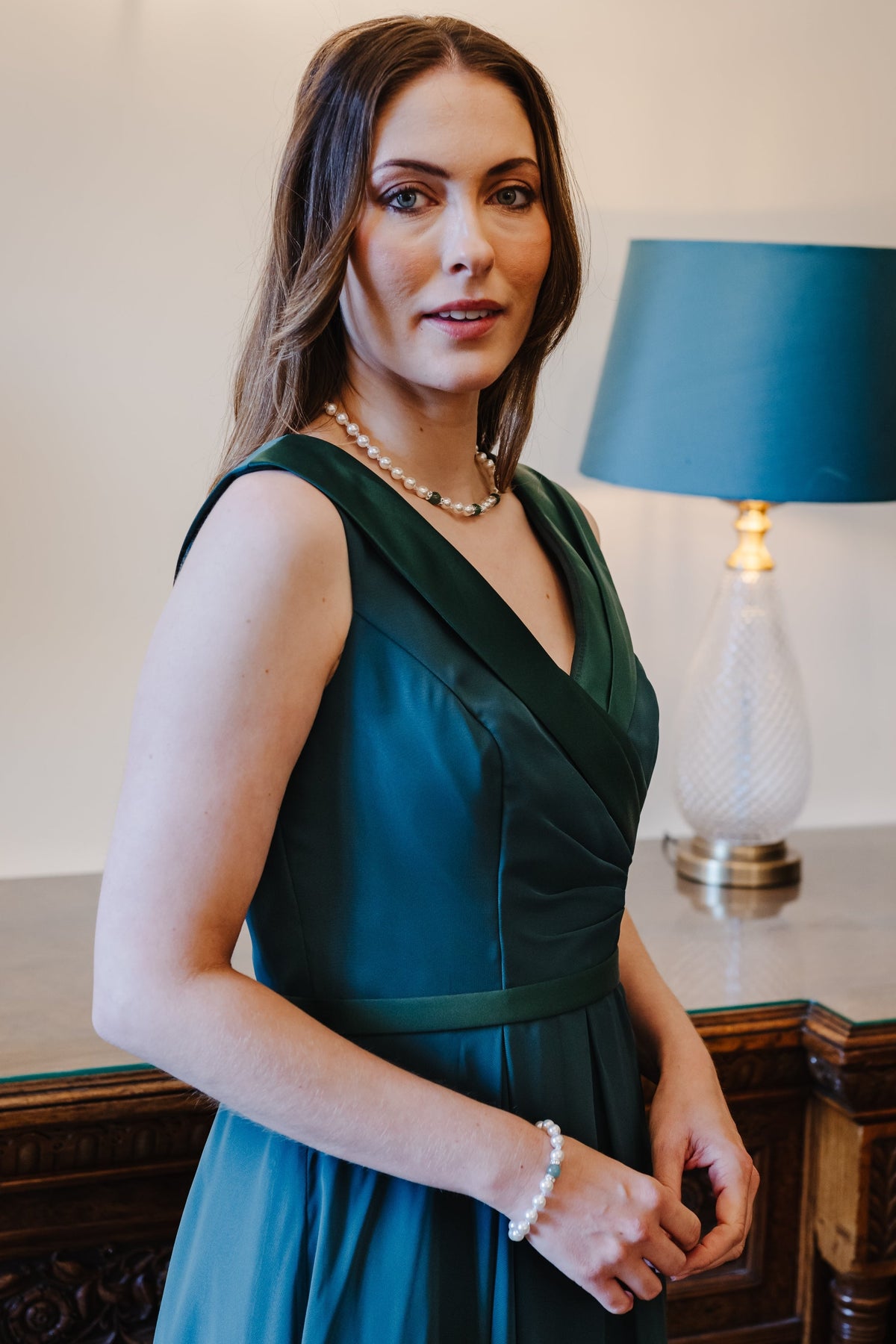 A woman wearing a sleeveless dark teal green wrap-style dress stands indoors in front of a lamp on a wooden table.