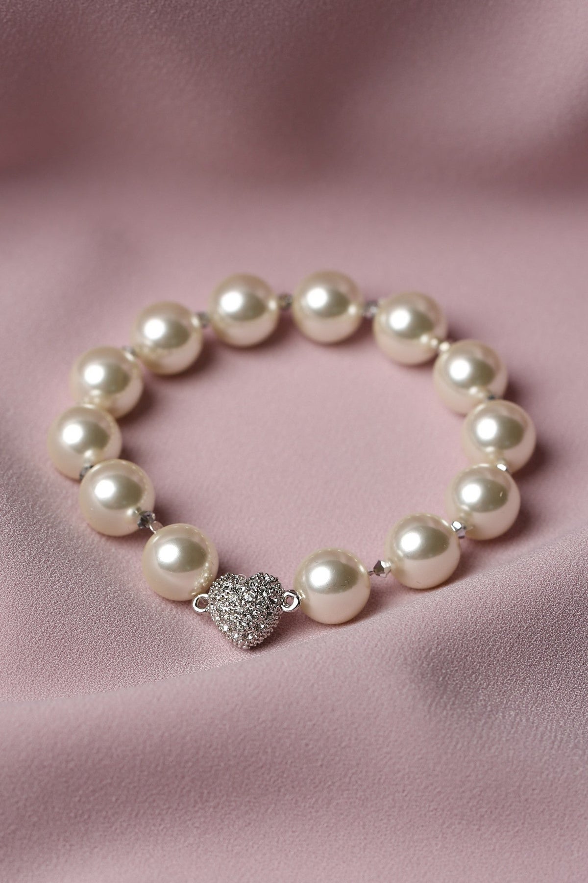 A pearl beaded bracelet with a silver heart-shaped charm encrusted with small clear stones, displayed on a soft pink satin fabric background.