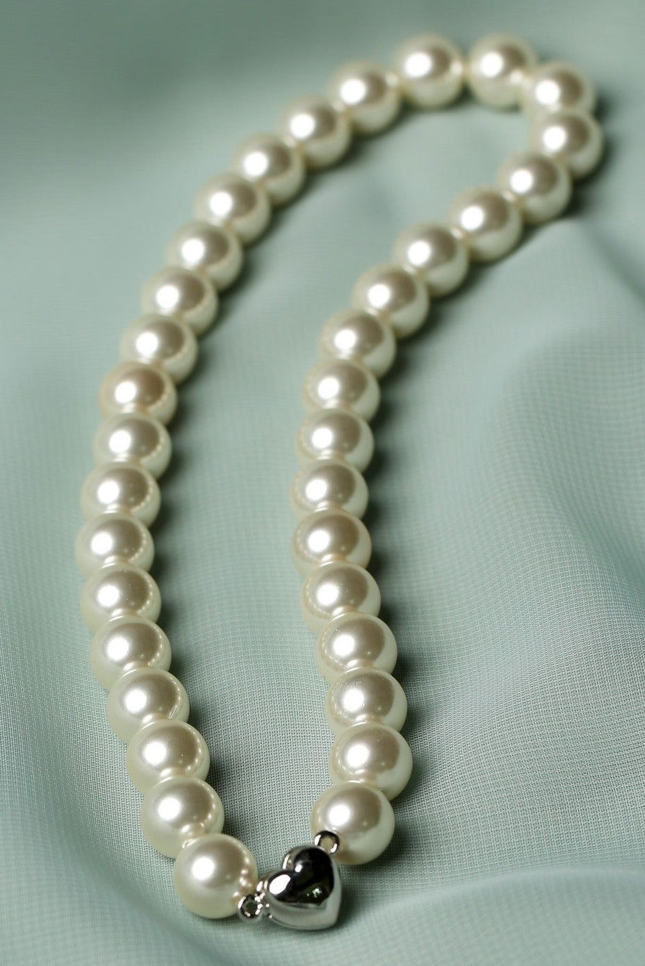 A strand of white freshwater pearls arranged in a loop on a light sage green satin fabric background, featuring a small silver heart-shaped charm at the end.