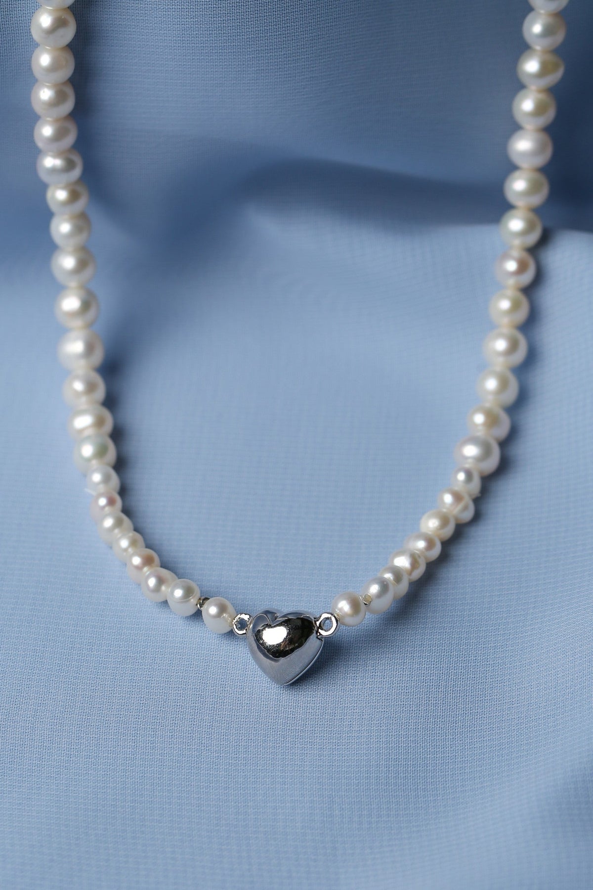 A pearl necklace with a polished silver heart pendant, displayed on a soft, draped light blue fabric background.