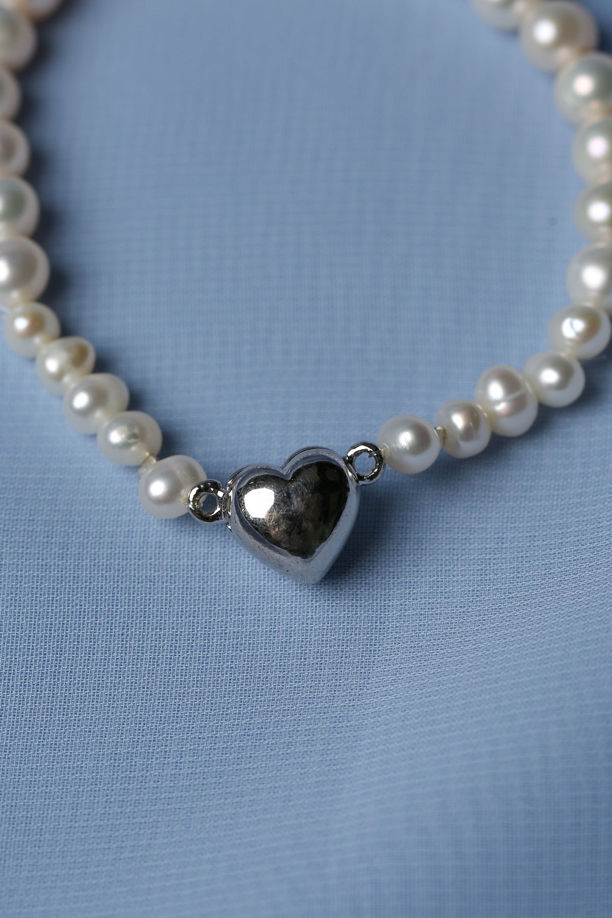 A close-up of a pearl strand necklace with a polished silver heart-shaped pendant, resting on a textured light blue fabric background.