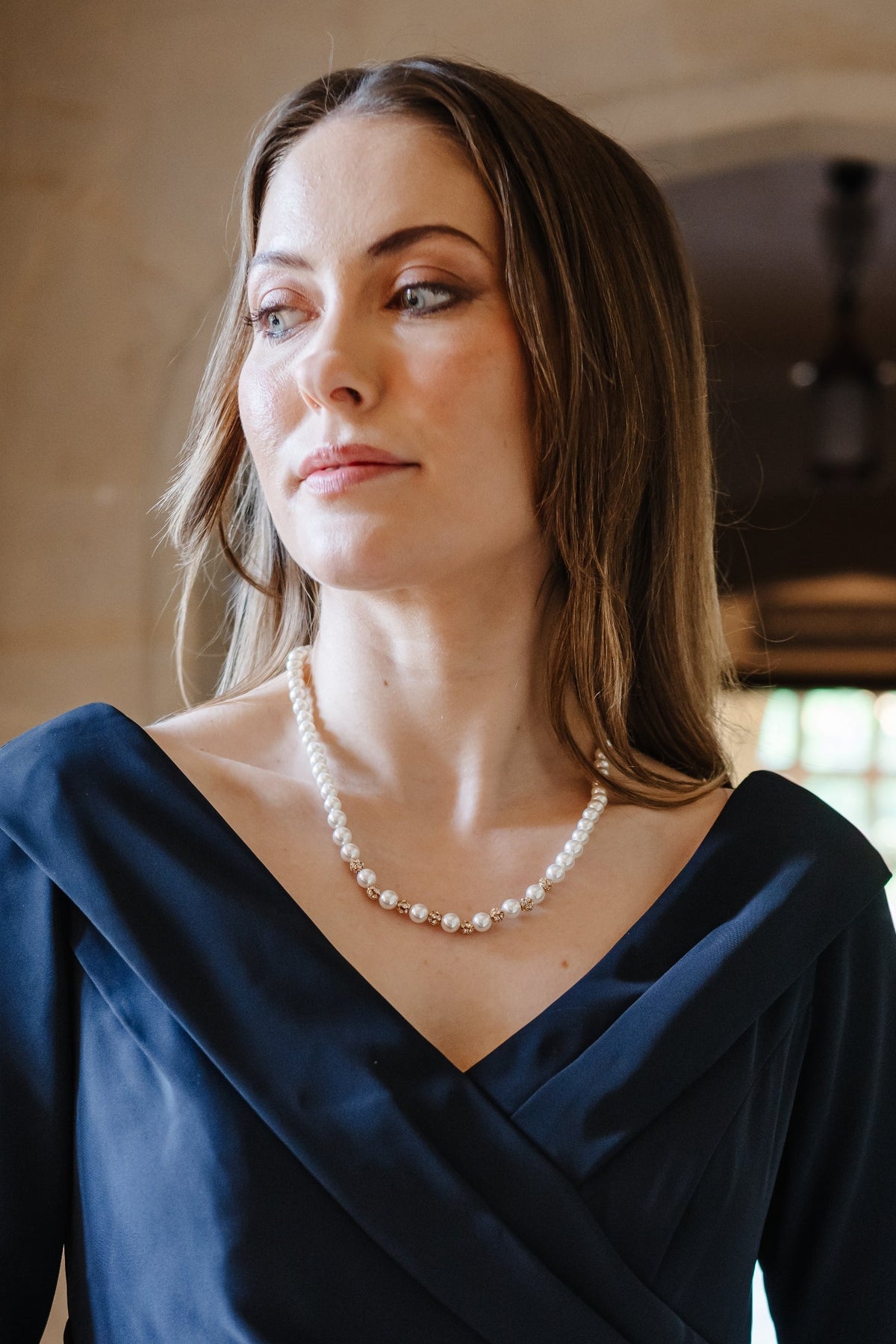 A woman wearing a navy blue wrap-style dress with three-quarter sleeves and a pearl necklace, standing in a softly lit indoor setting with stone arches and a blurred background.