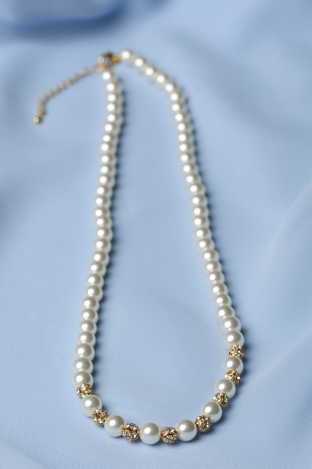 A pearl necklace with gold floral accents and a gold clasp, displayed on a soft light blue fabric background.