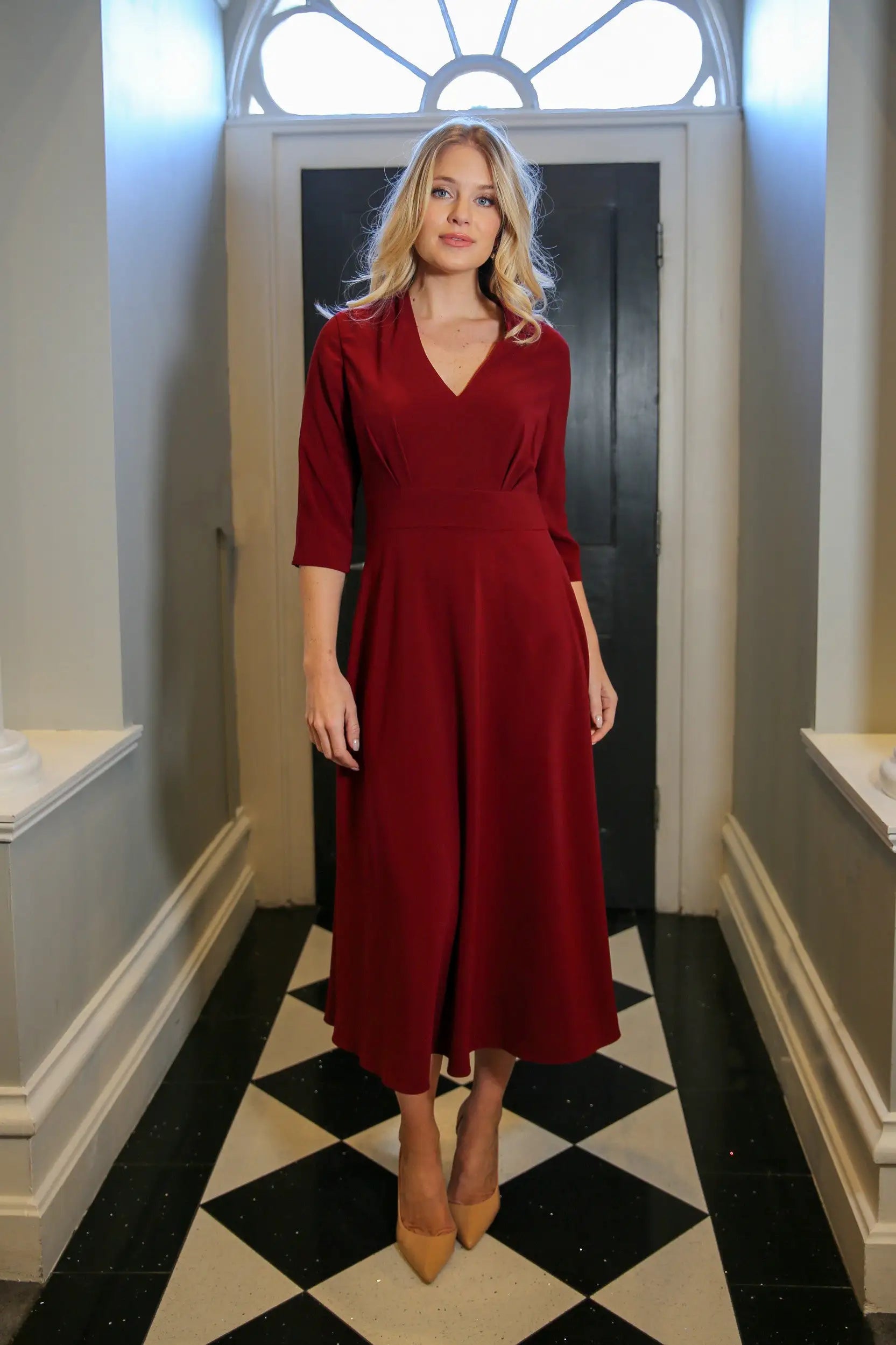 A model wearing a red, midi-length dress with a V-neck and 3/4 sleeves, standing in a room with black and white tiled floor.