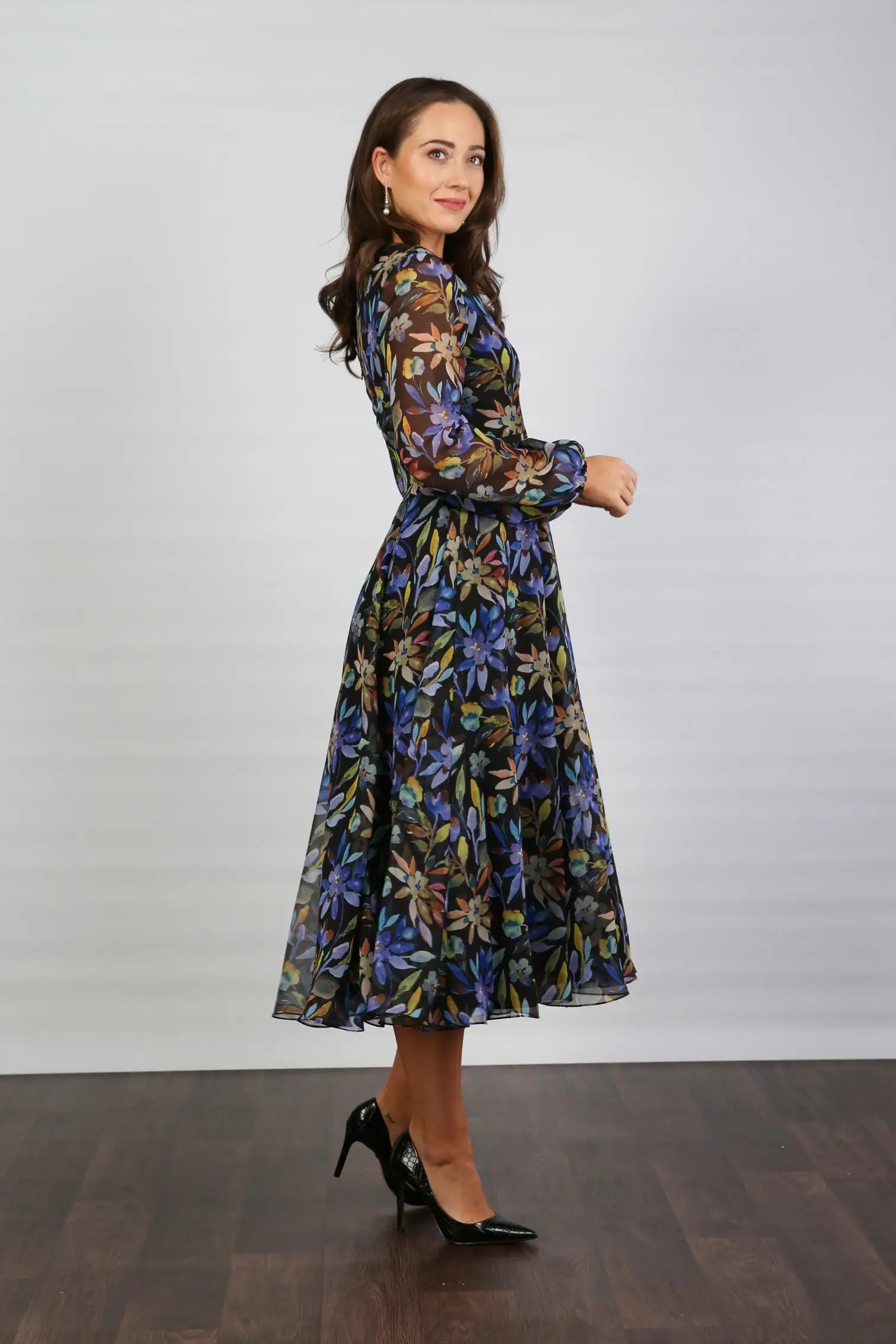 A woman standing indoors wearing a floral printed chiffon midi dress with long sleeves and a round neckline.