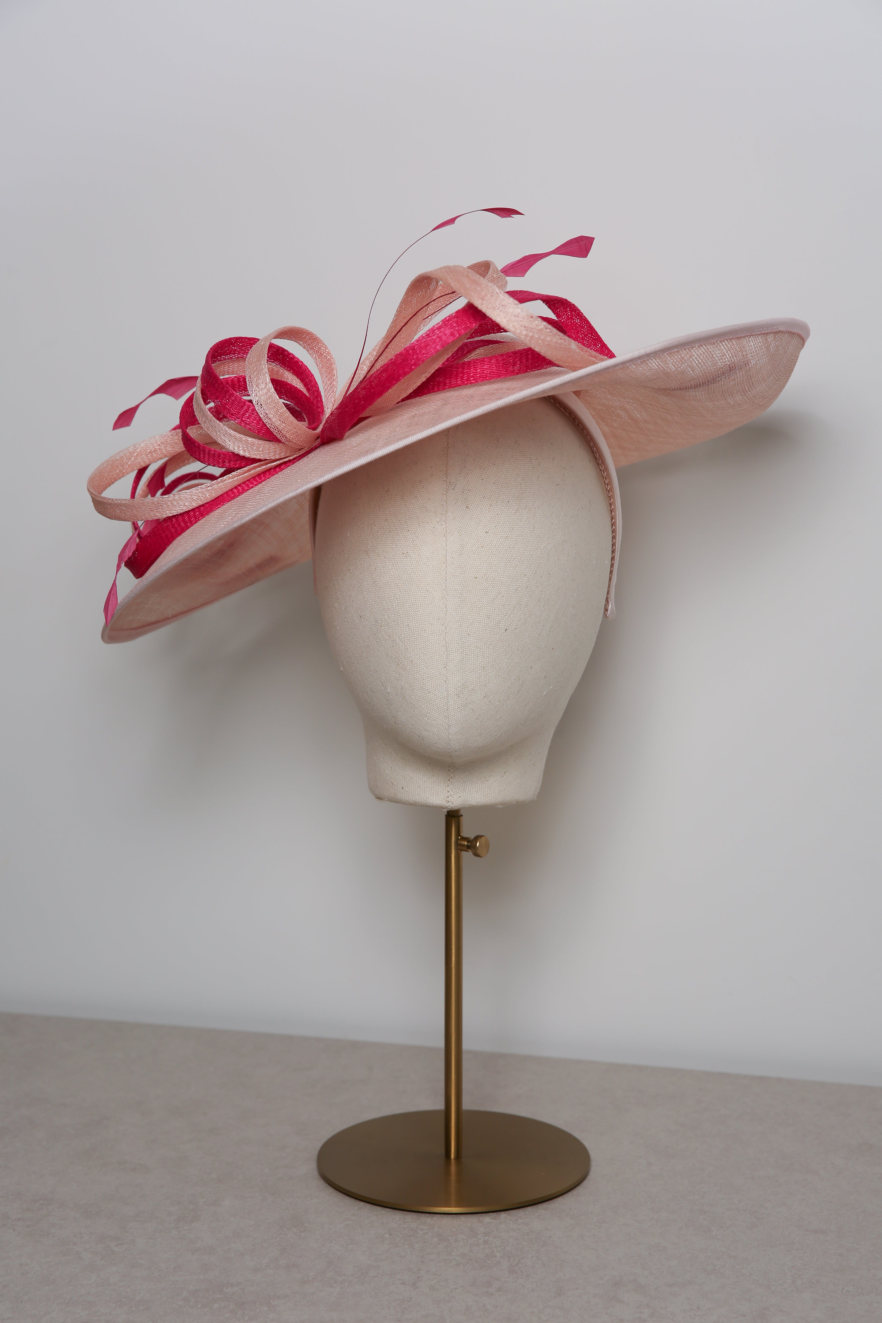 Pink and white decorative hat on a mannequin head with a gold stand against a plain background