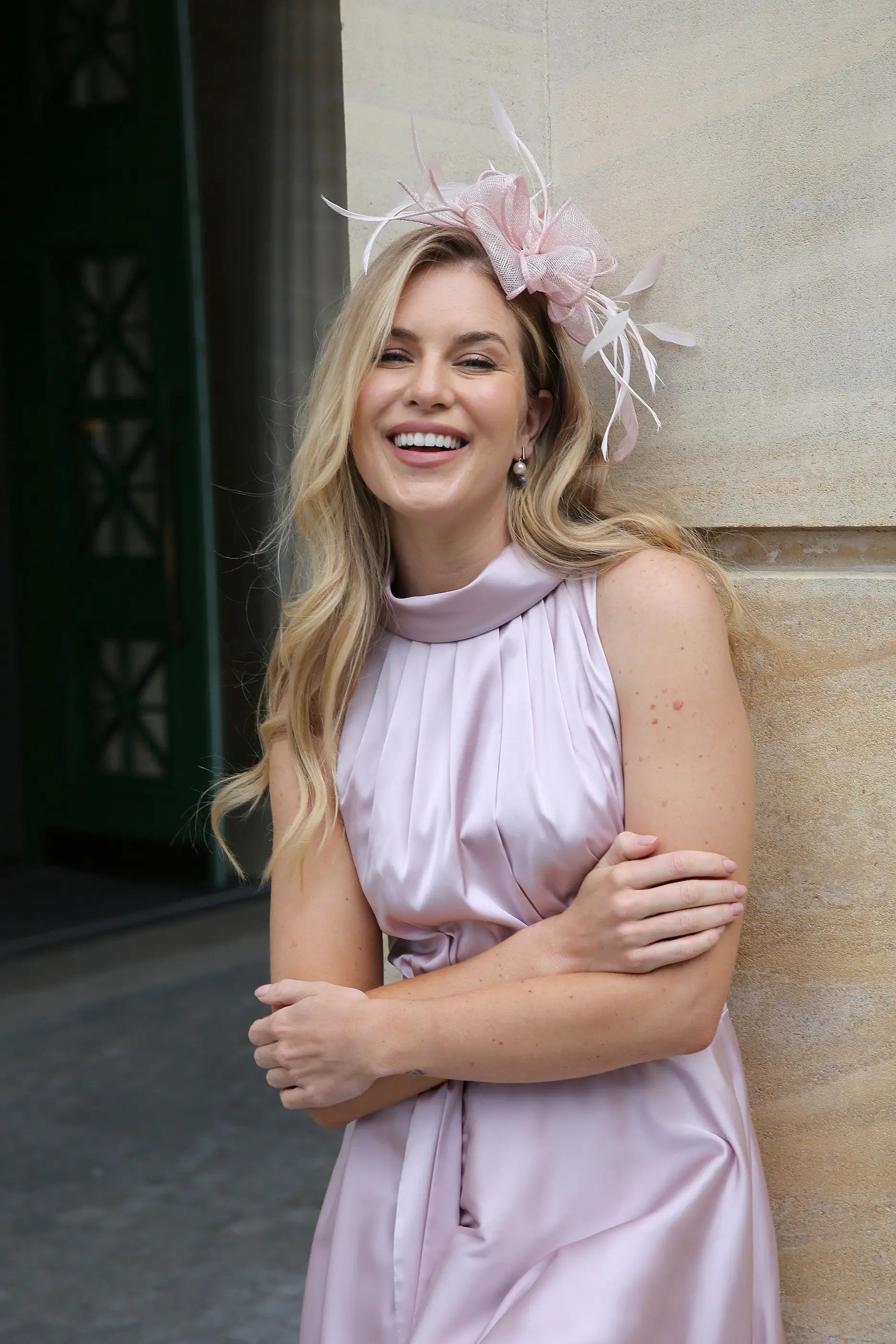 A woman wearing a pink sleeveless satin dress with a high neck and self-tie waistband, standing in front of a building. Styled with a matching fascinator.