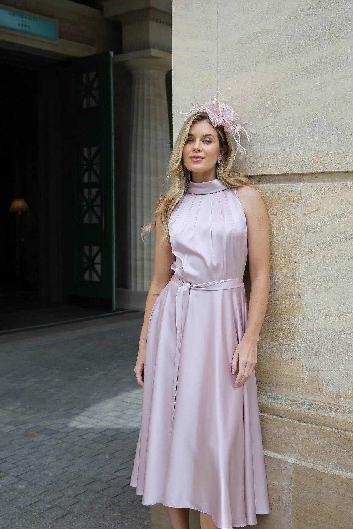 A woman wearing a pink sleeveless satin dress with a high neck and self-tie waistband, standing in front of a building. Styled with a matching fascinator.