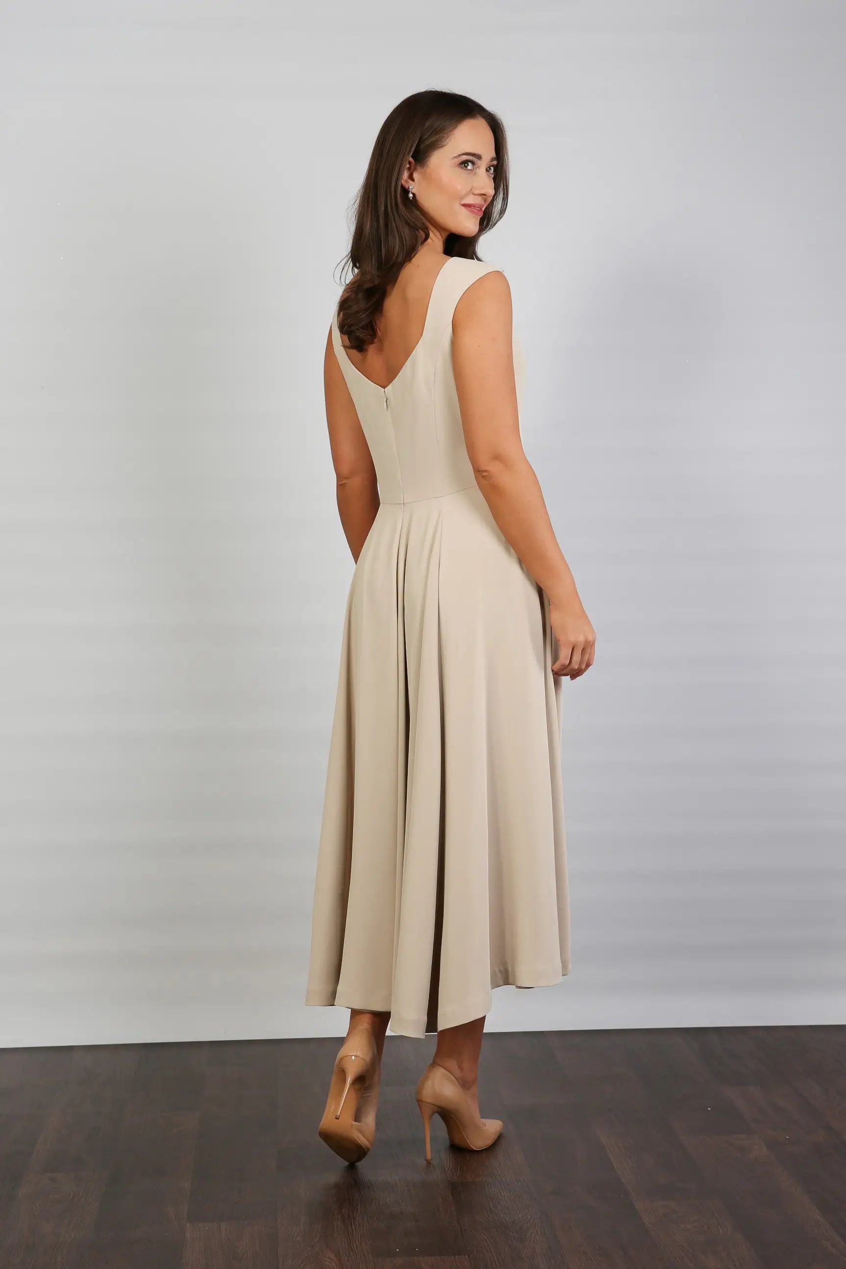 A woman standing in a semi-formal, sleeveless, midi length beige dress with a round neckline and a full skirt.