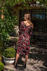Woman in a burgundy floral dress standing outdoors with greenery and a building in the background.