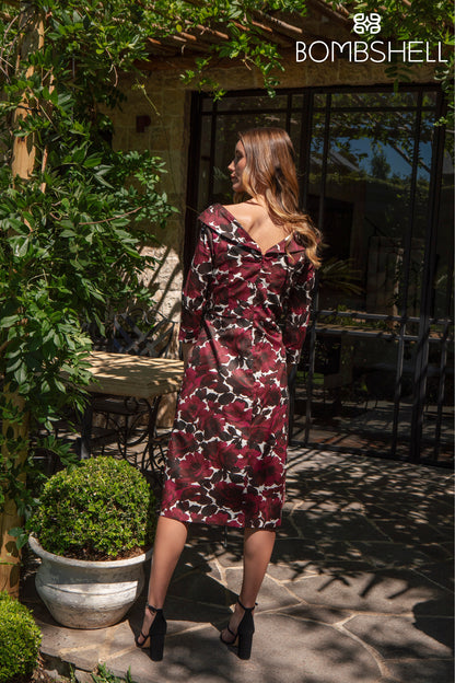 Woman in a burgundy floral dress standing outdoors with greenery and a building in the background.