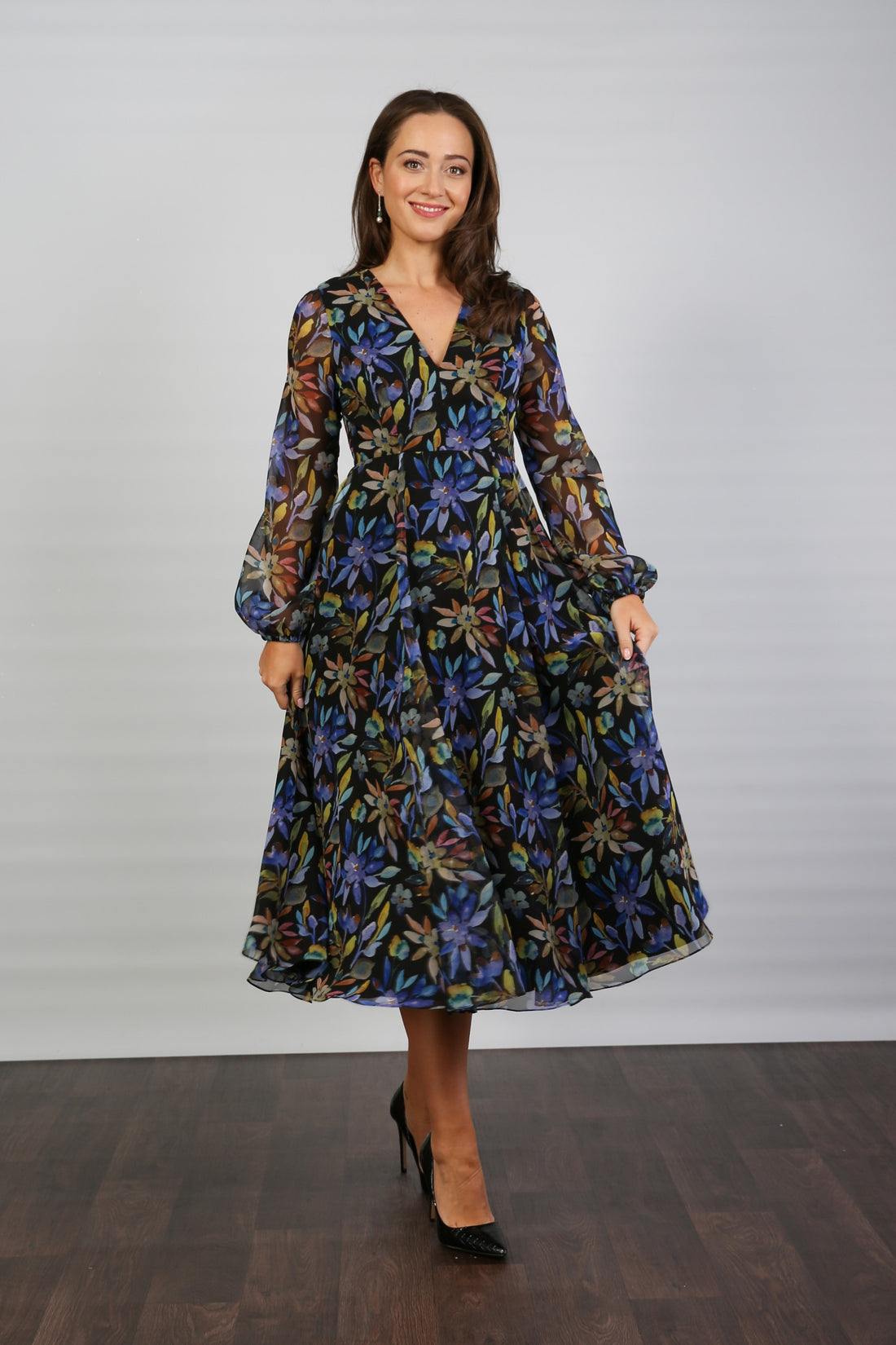 A woman standing indoors wearing a floral printed chiffon midi dress with long sleeves and a round neckline.