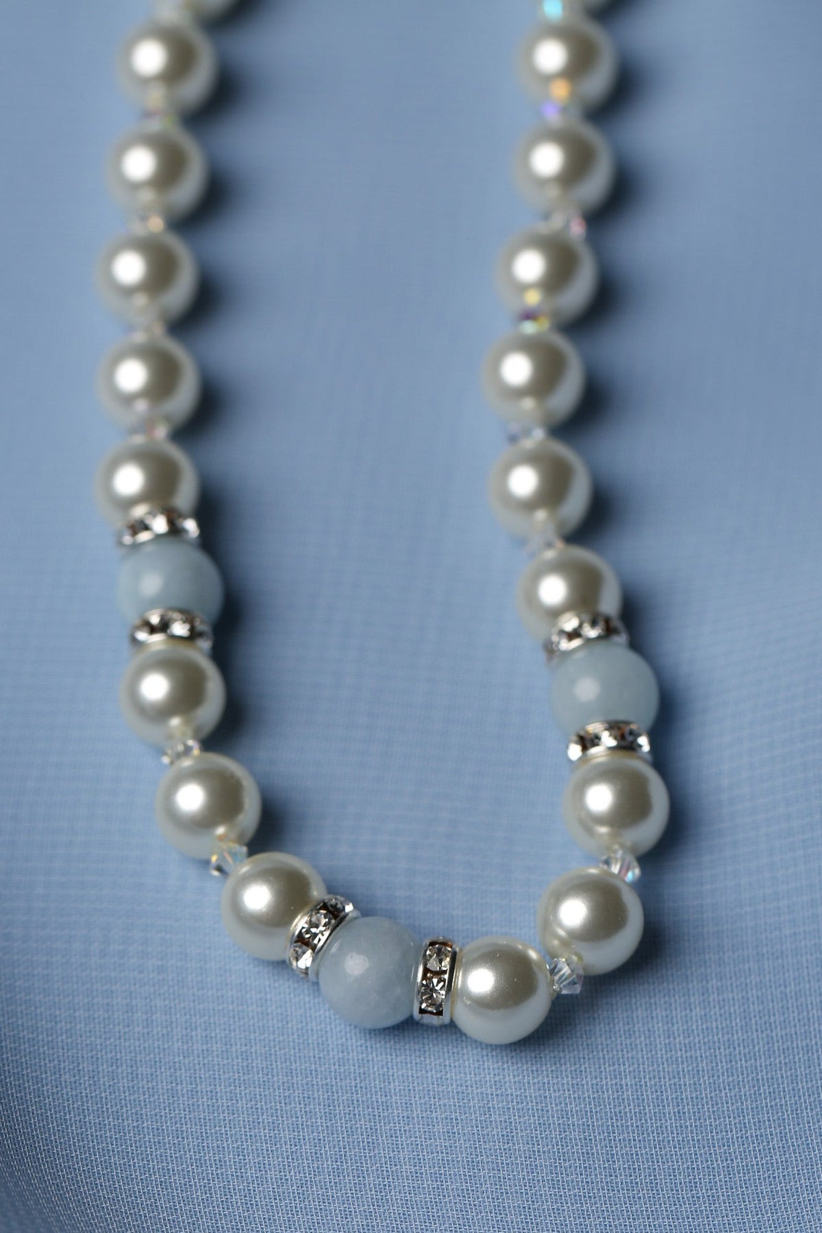 A beaded necklace featuring white pearl-like round beads, light blue stone beads, and small silver spacer rings, laid on a soft light blue fabric background.