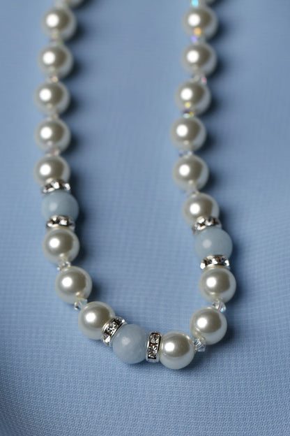 A beaded necklace featuring white pearl-like round beads, light blue stone beads, and small silver spacer rings, laid on a soft light blue fabric background.