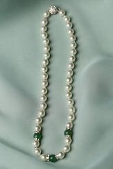 A pearl necklace with small green stone accents and silver-toned spacers, laid flat on a soft pale green satin fabric background.