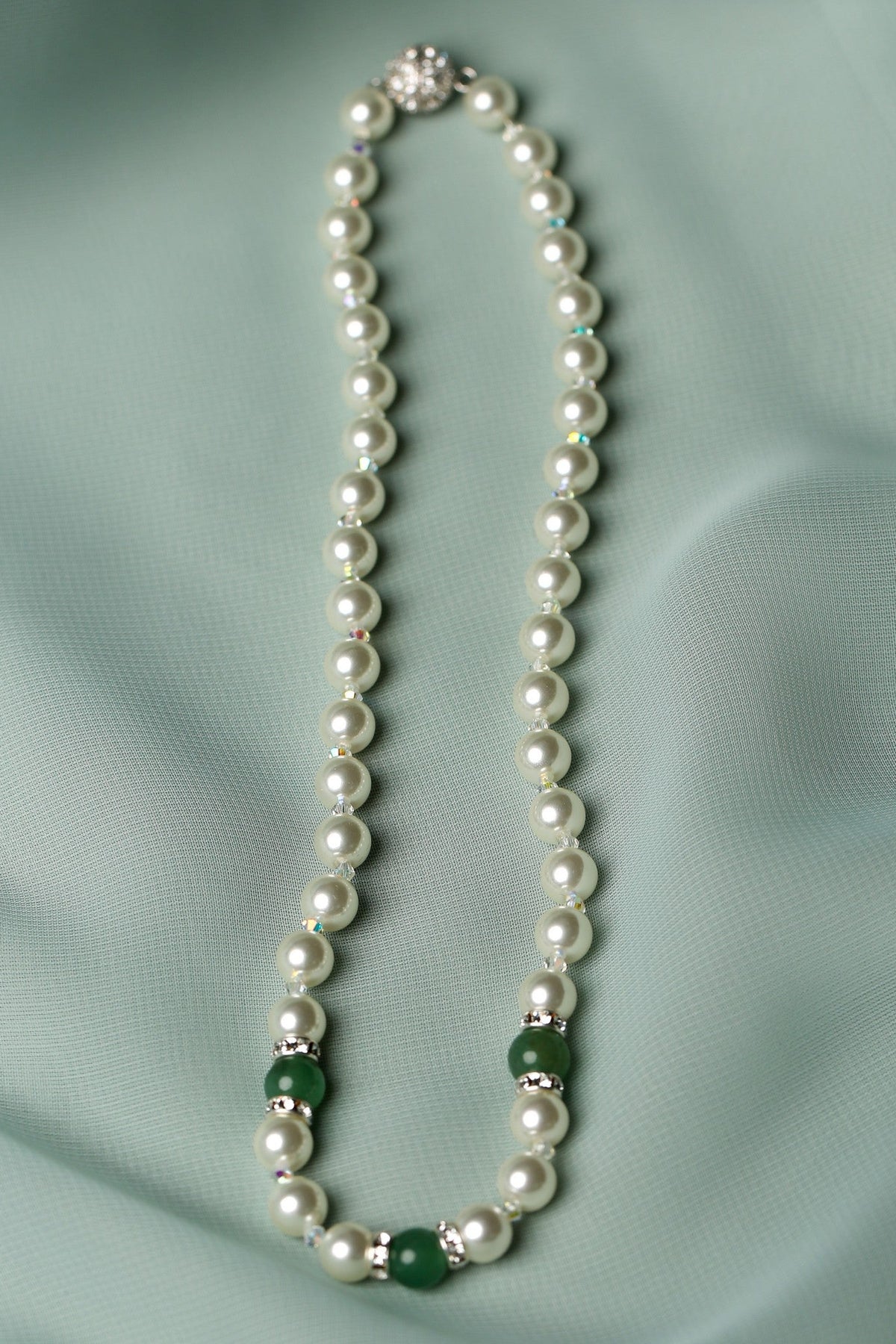 A pearl necklace with small green stone accents and silver-toned spacers, laid flat on a soft pale green satin fabric background.
