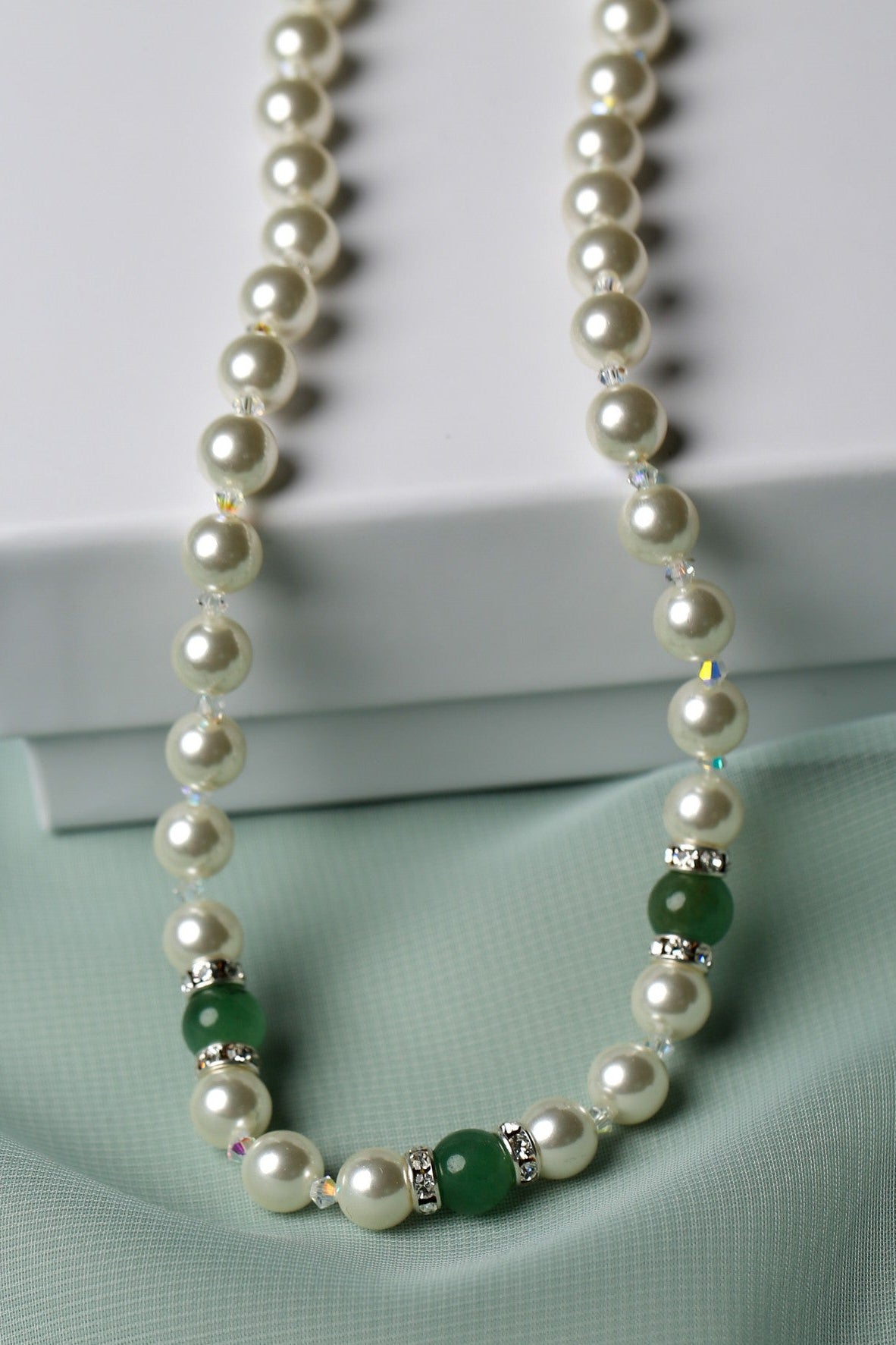 A pearl necklace with green jade-like beads and silver spacer accents, draped elegantly over a white box on a soft mint green fabric background.