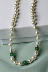 A pearl necklace with green jade-like beads and silver spacer accents, draped elegantly over a white box on a soft mint green fabric background.