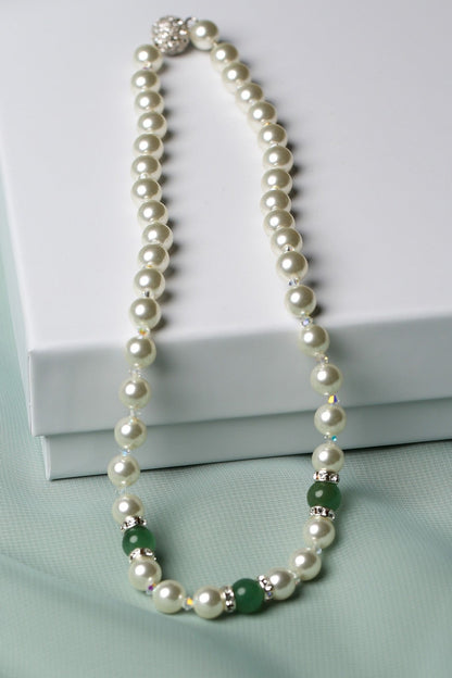 A pearl necklace with small green and silver accent beads, draped elegantly over a white gift box on a soft mint green fabric background.