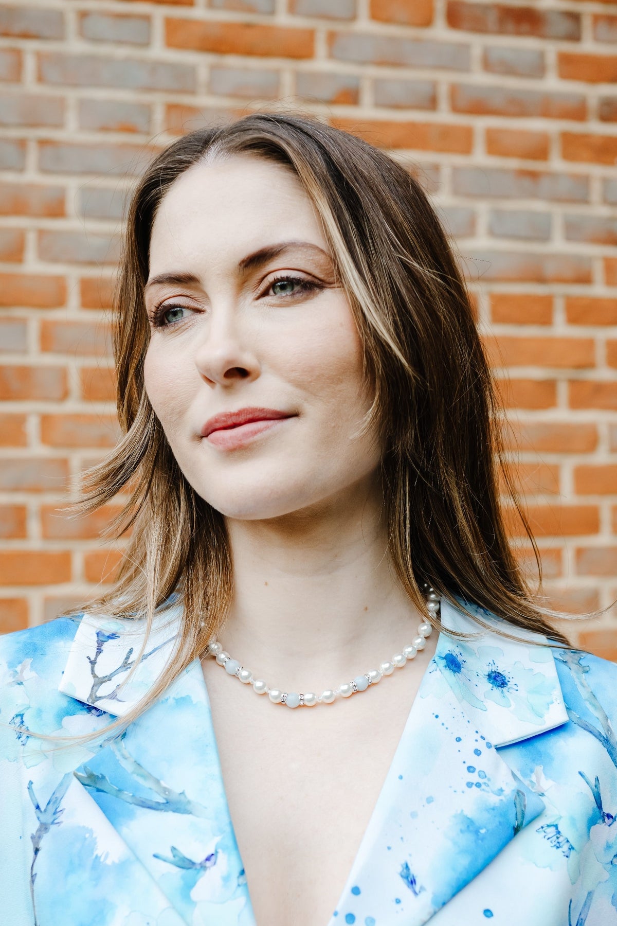 A woman with brown hair wearing a light blue watercolor floral print blazer and a pearl necklace, standing in front of a red brick wall.
