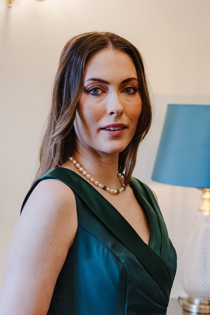 A woman wearing a sleeveless dark teal green wrap-style dress stands indoors in front of a lamp on a wooden table.