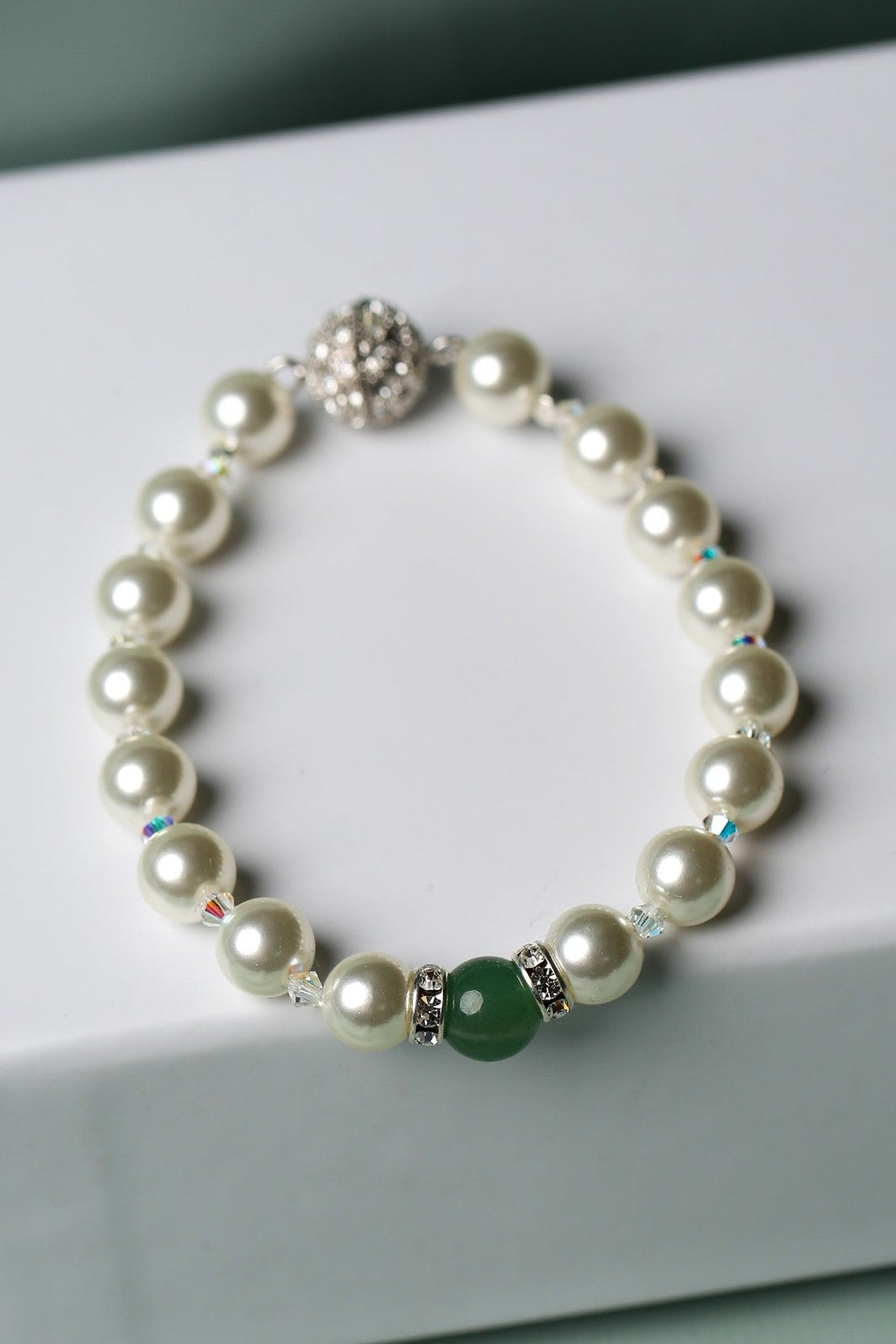 A pearl beaded bracelet with a green round bead and silver-toned metal accents, displayed on a white gift box against a soft mint green background.