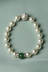 A pearl beaded bracelet with a silver-toned clasp, featuring a single green stone accent bead, displayed on a soft sage green fabric background.