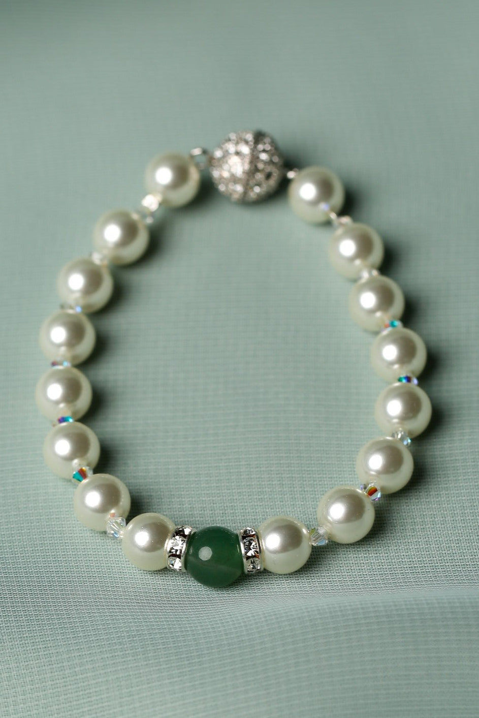A pearl beaded bracelet with a silver-toned clasp, featuring a single green stone accent bead, displayed on a soft sage green fabric background.