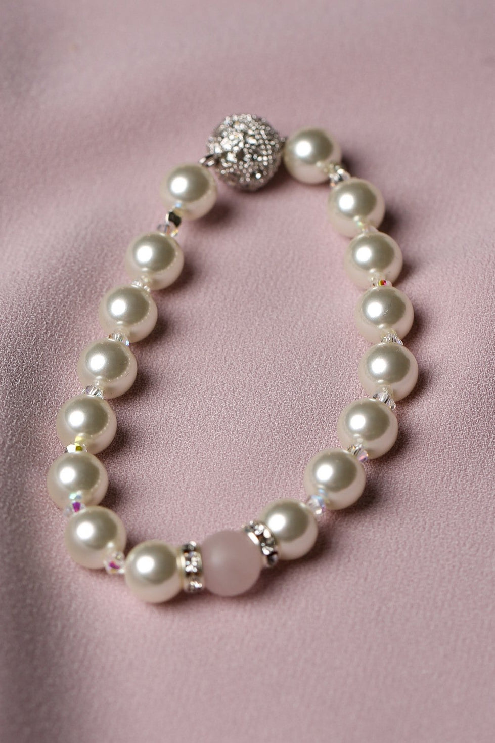 A pearl beaded bracelet with a silver-toned round clasp and a pink accent bead, displayed on a soft pink satin fabric background.