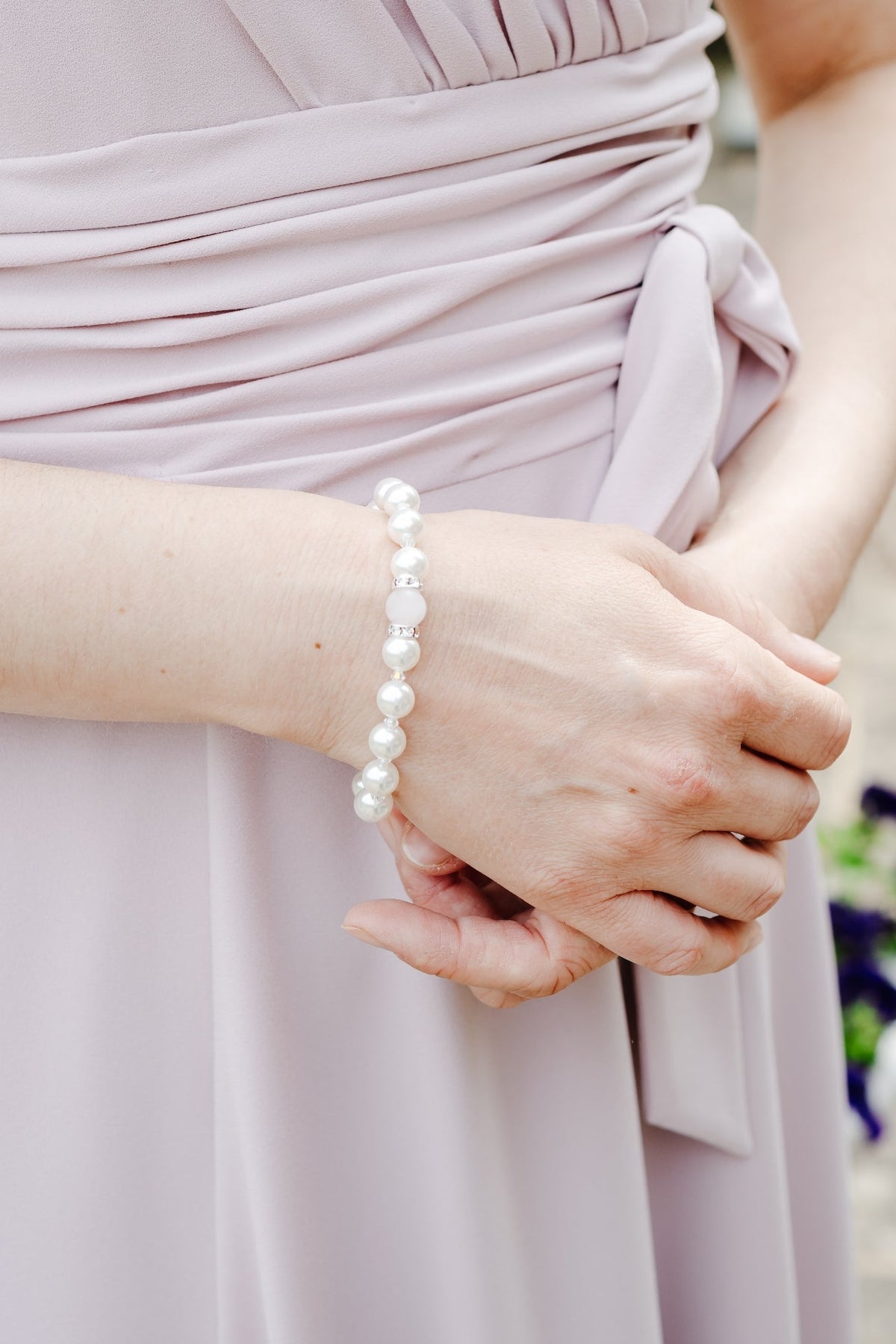 A close-up of a person wearing a light lavender wrap-style dress with a tied waist sash, shown outdoors with hands clasped and a pearl bracelet visible on one wrist.