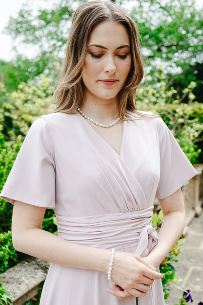 A woman wearing a light lavender wrap-style dress with short sleeves and a tied waist, standing outdoors in a garden setting with greenery and stone steps in the background.