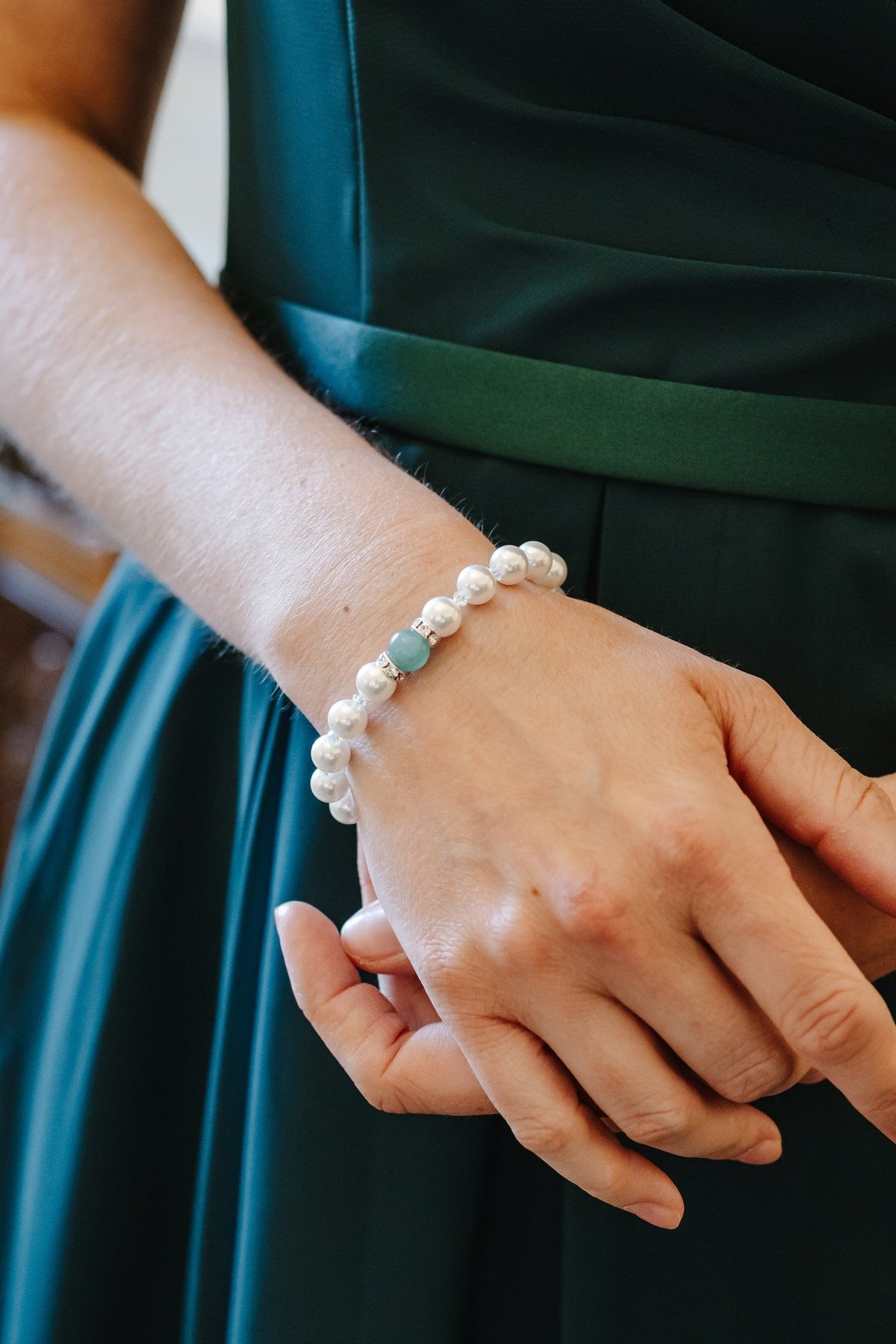 A close-up of a person wearing a dark teal green satin dress with a matching waistband, showcasing a white pearl beaded bracelet with a small green stone on their wrist.