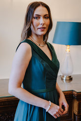 A woman wearing a sleeveless dark teal green wrap-style dress stands indoors in front of a lamp on a wooden table.