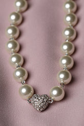 A pearl necklace with a silver heart-shaped pavé centerpiece, displayed on a soft pink satin fabric background.