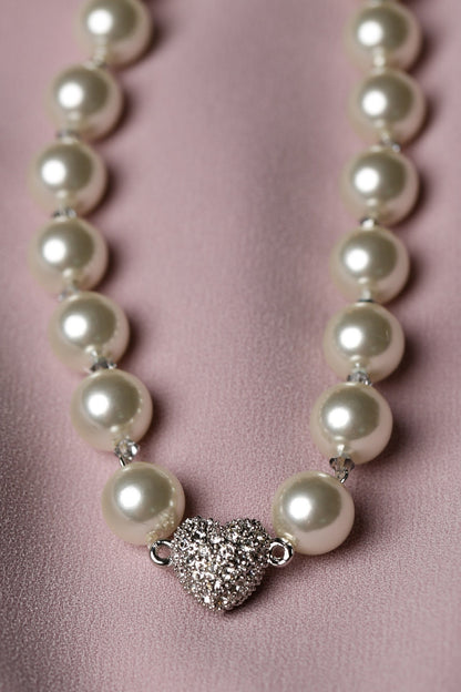A pearl necklace with a silver heart-shaped pavé centerpiece, displayed on a soft pink satin fabric background.