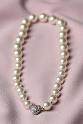 A strand of white faux-pearl beads arranged in a circle on a soft pink satin fabric background, featuring a silver-toned heart-shaped charm embellished with small clear rhinestones at the bottom center.