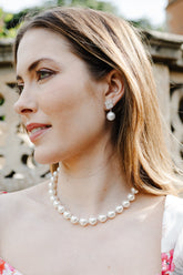 A woman with blonde hair is shown in profile wearing a pearl necklace and matching pearl drop earrings, with a floral white top and a stone garden wall in the background.