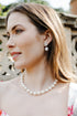 A woman with blonde hair is shown in profile wearing a pearl necklace and matching pearl drop earrings, with a floral white top and a stone garden wall in the background.