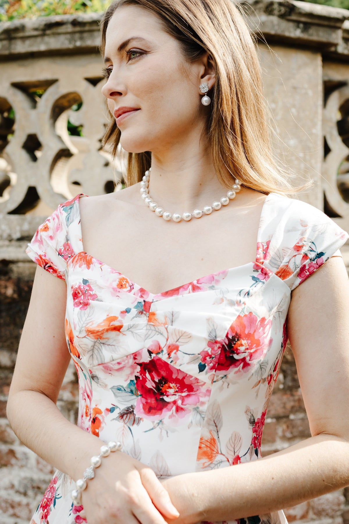 A woman wearing a white floral dress with red and orange flowers, accessorized with a pearl necklace and bracelet, standing outdoors in front of a stone wall.