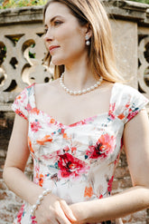 A woman wearing a white floral dress with red and orange flowers, accessorized with a pearl necklace and bracelet, standing outdoors in front of a stone wall.