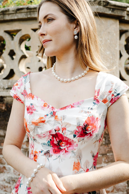 A woman wearing a white floral dress with red and orange flowers, accessorized with a pearl necklace and bracelet, standing outdoors in front of a stone wall.