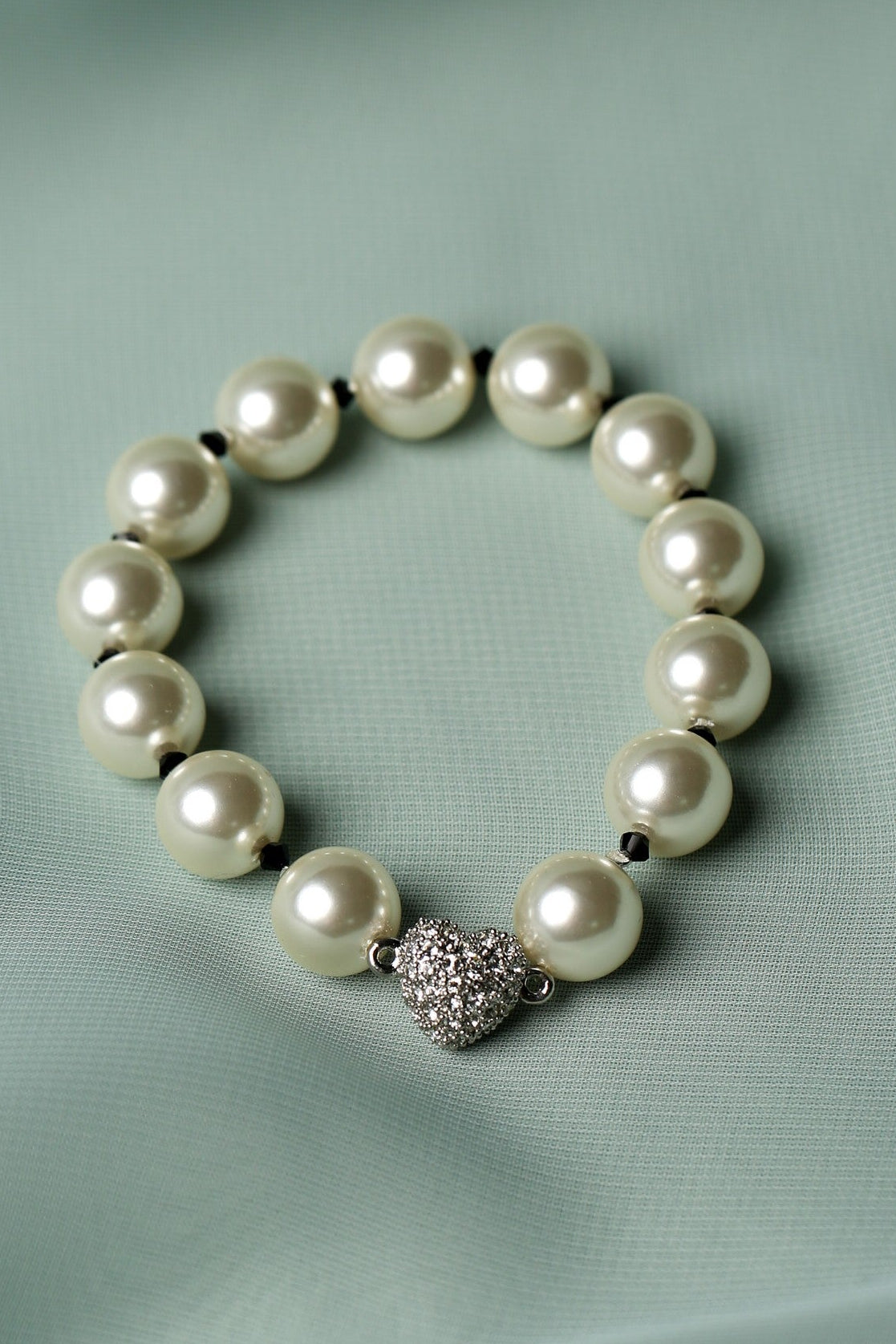 A pearl beaded bracelet with a textured silver heart charm, displayed on a soft, light green fabric background.