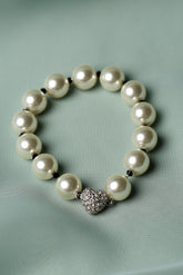 A pearl beaded bracelet with a textured silver heart charm, displayed on a soft, light green fabric background.