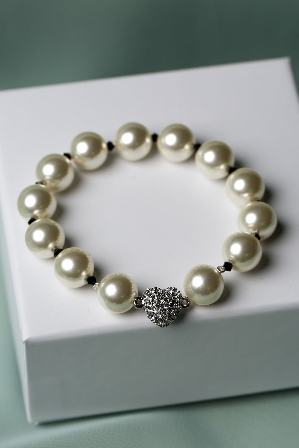 A pearl beaded bracelet with a silver pavé heart charm, displayed on a white gift box against a soft sage green background.