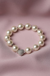 A pearl beaded bracelet with a silver heart-shaped charm encrusted with small clear stones, displayed on a soft pink satin fabric background.