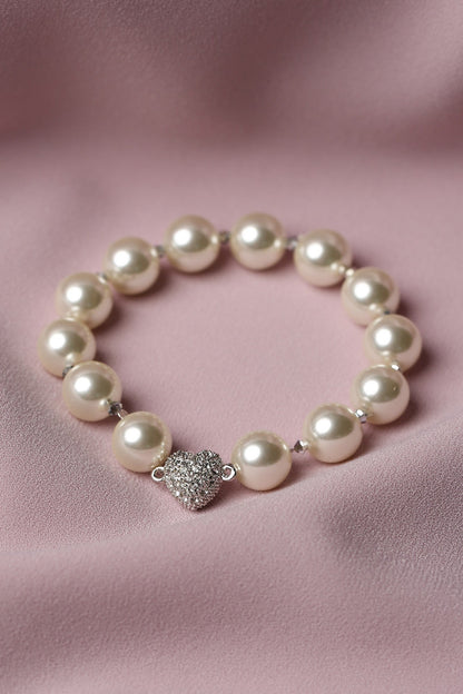 A pearl beaded bracelet with a silver heart-shaped charm encrusted with small clear stones, displayed on a soft pink satin fabric background.