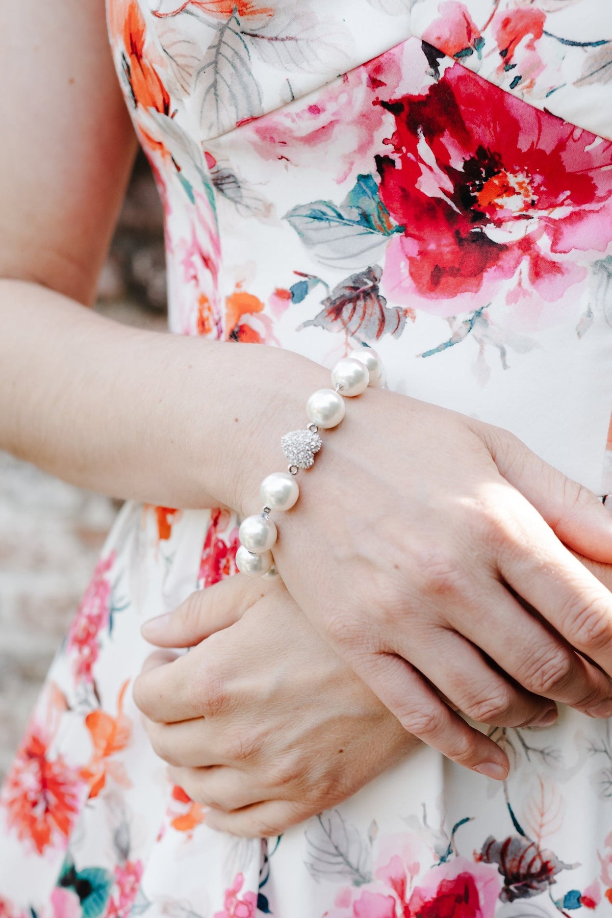 A close-up of a woman wearing a white sleeveless dress with a vibrant red, pink, and orange floral print. She is wearing a pearl bracelet on her wrist, with her hands crossed over her waist, set against a blurred brick wall background.